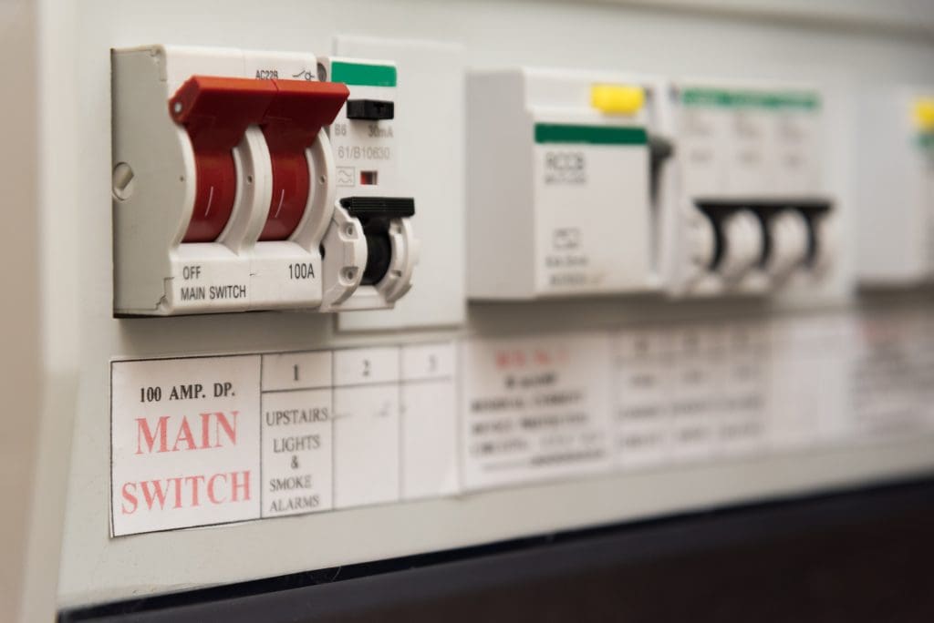 Close-up of an electrical panel with switches and labels. The red switch labeled "MAIN SWITCH" is prominent, and there are other switches with white toggles and labels indicating functions like "UPSTAIRS LIGHTS & SMOKE ALARMS.