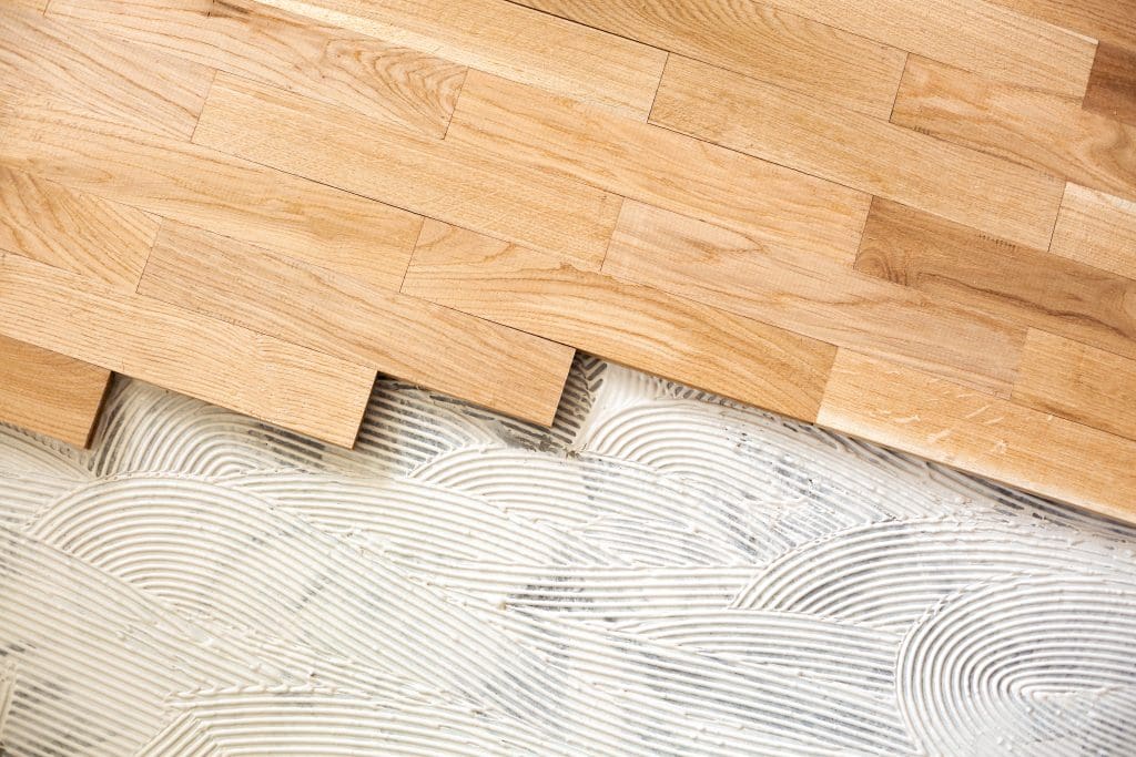 Close-up of a partially installed hardwood floor. The natural wood planks are placed over a surface with visible adhesive in wavy patterns. The upper part shows the finished flooring, while the lower part is incomplete.