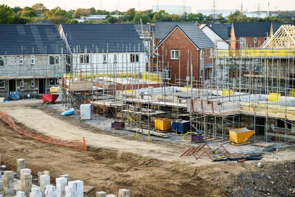 A construction site with partially built houses, surrounded by scaffolding and various construction materials. In the background, completed houses with grey and red roofs can be seen, along with trees and industrial buildings.