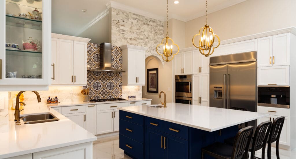 A modern kitchen featuring white cabinets, a navy island with black chairs, and a tiled backsplash. Two gold pendant lights hang above the island. Stainless steel appliances are visible, including a refrigerator and oven. A display cabinet holds teacups.