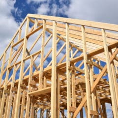 Wooden frame of a house under construction, set against a bright blue sky with scattered clouds. The beams and trusses are exposed, showcasing the skeleton of the structure.