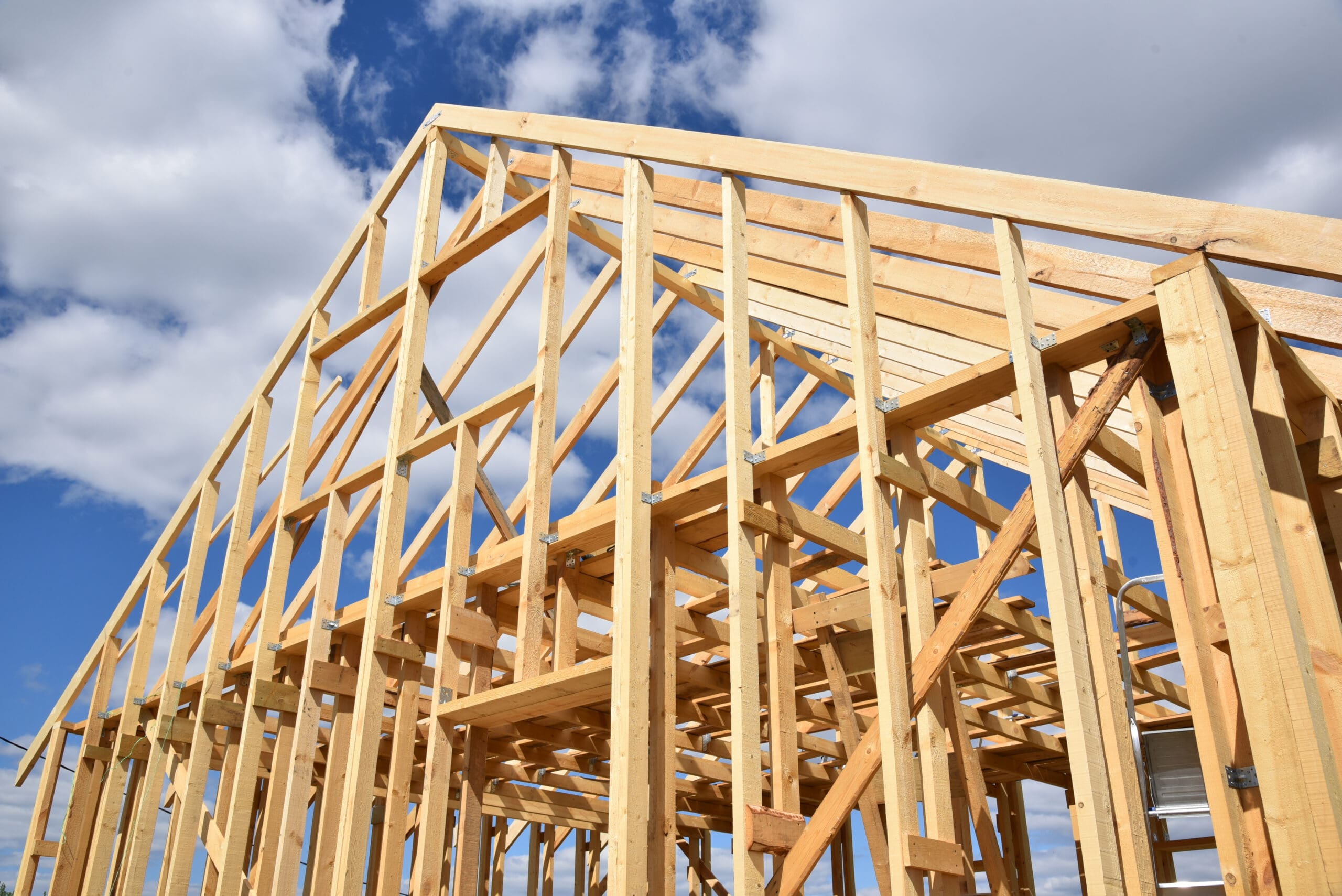 Wooden frame of a house under construction, set against a bright blue sky with scattered clouds. The beams and trusses are exposed, showcasing the skeleton of the structure.