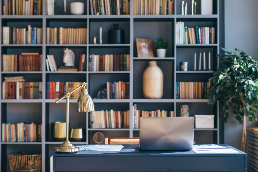 A modern home office setup with a laptop on a sleek black desk. Behind it, a large bookshelf filled with various books and decorative items, including a potted plant, vases, and sculptures. A brass desk lamp adds elegance to the workspace.