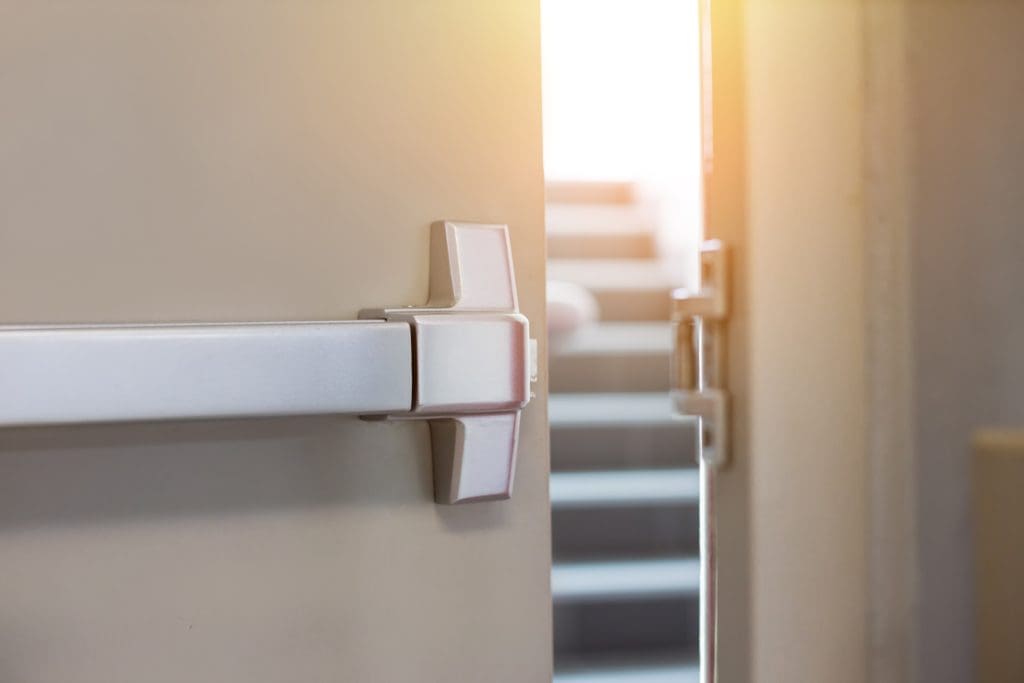 A door with a metal push bar is slightly open, leading to a staircase. Sunlight is streaming in from the top of the stairs, creating a bright and warm atmosphere in the otherwise dimly lit area.