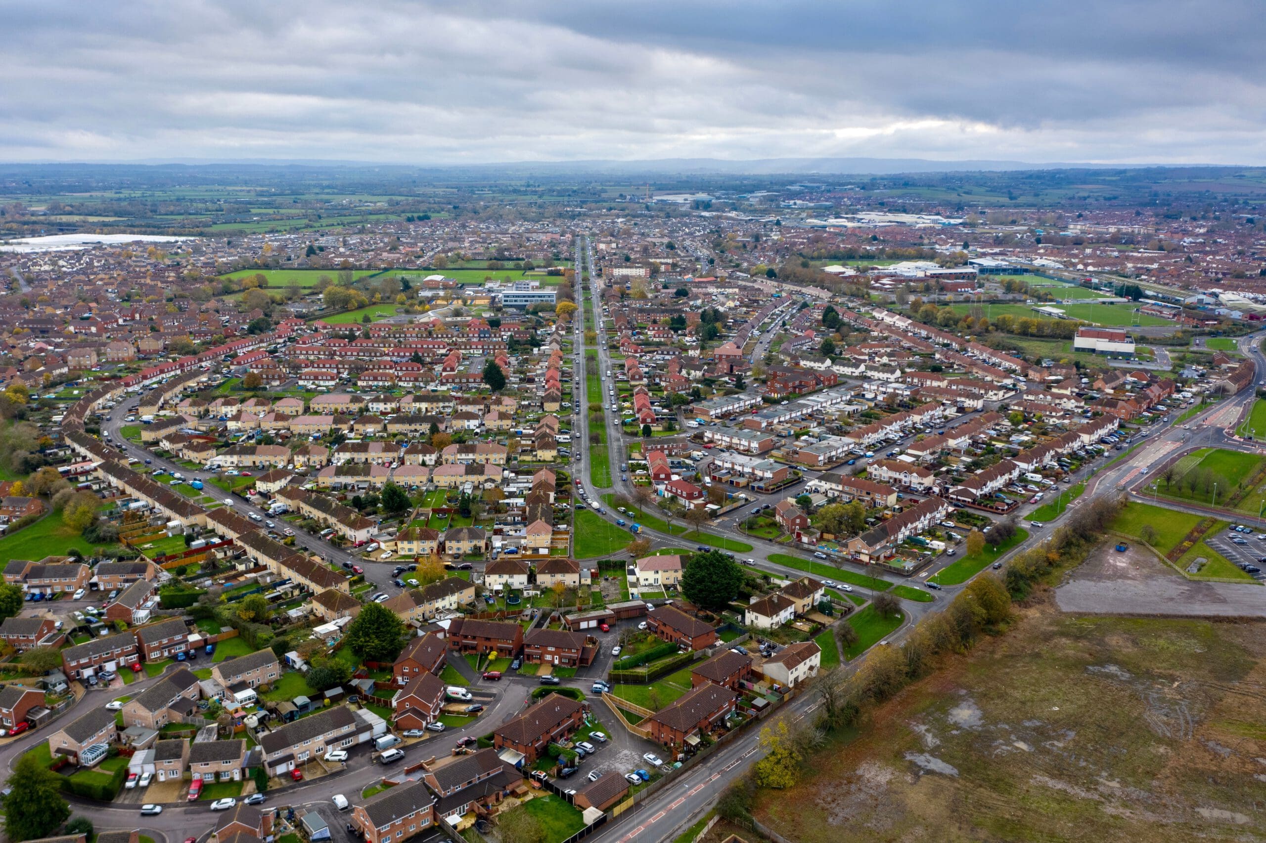 Aerial view of a sprawling suburban neighborhood with rows of houses, intersecting roads, and patches of greenery. The landscape extends into the distance under a cloudy sky.