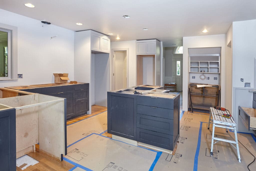 A modern kitchen under renovation with dark cabinets, recessed lighting, and wood flooring. The room is unfurnished, with construction materials and equipment visible. Walls are white, and cardboard sheets protect the floor.