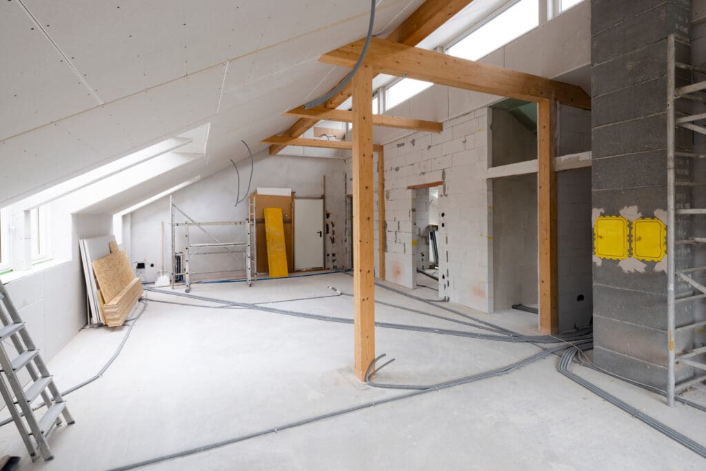 An unfinished attic space with exposed wooden beams, insulation panels, and scattered electrical wires. Scaffolding, ladders, and construction materials are visible, with partially constructed walls and ceiling under natural light from skylights.