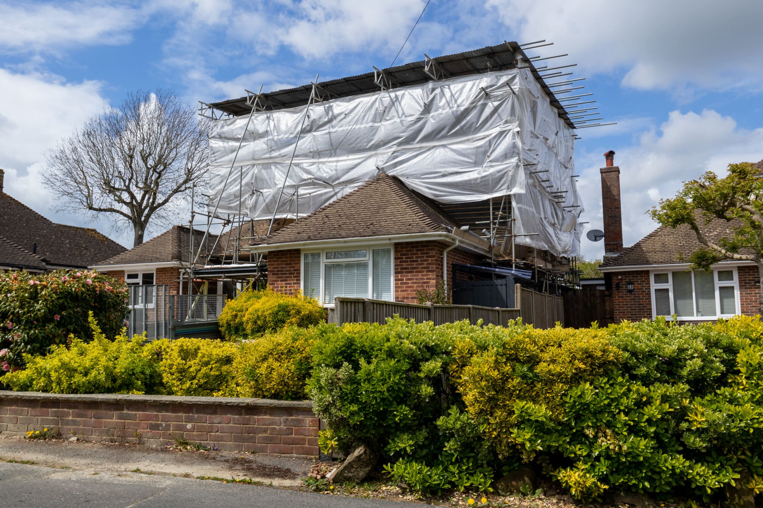 A brick house with a large temporary structure covered in white tarps on the roof. Scaffolding surrounds the upper portion, extending into the sky. There's a hedge and a bare tree in the foreground under a partly cloudy sky.