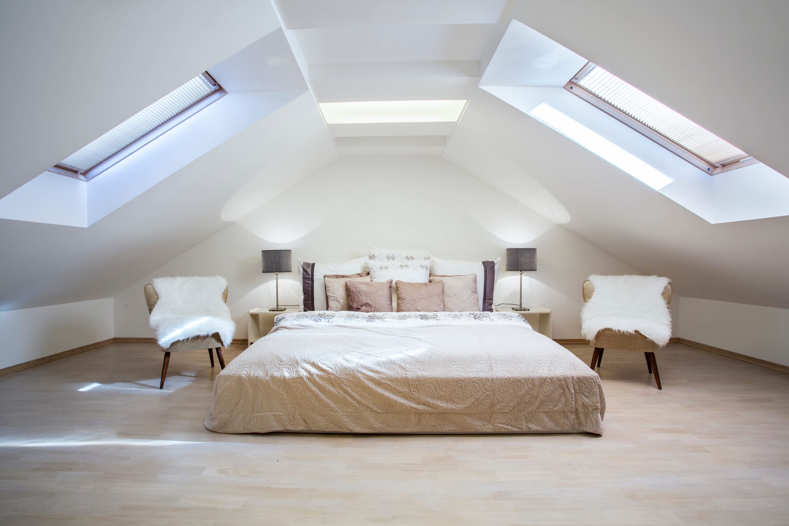 A cozy attic bedroom with a large bed covered in beige bedding. Two chairs with fur cushions flank the bed. The room has a wooden floor and is illuminated by skylights, creating a bright and airy atmosphere.