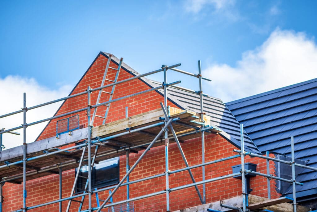 A brick house under construction with scaffolding surrounding it. The roof is partially completed with dark gray tiles. The sky is clear and blue.