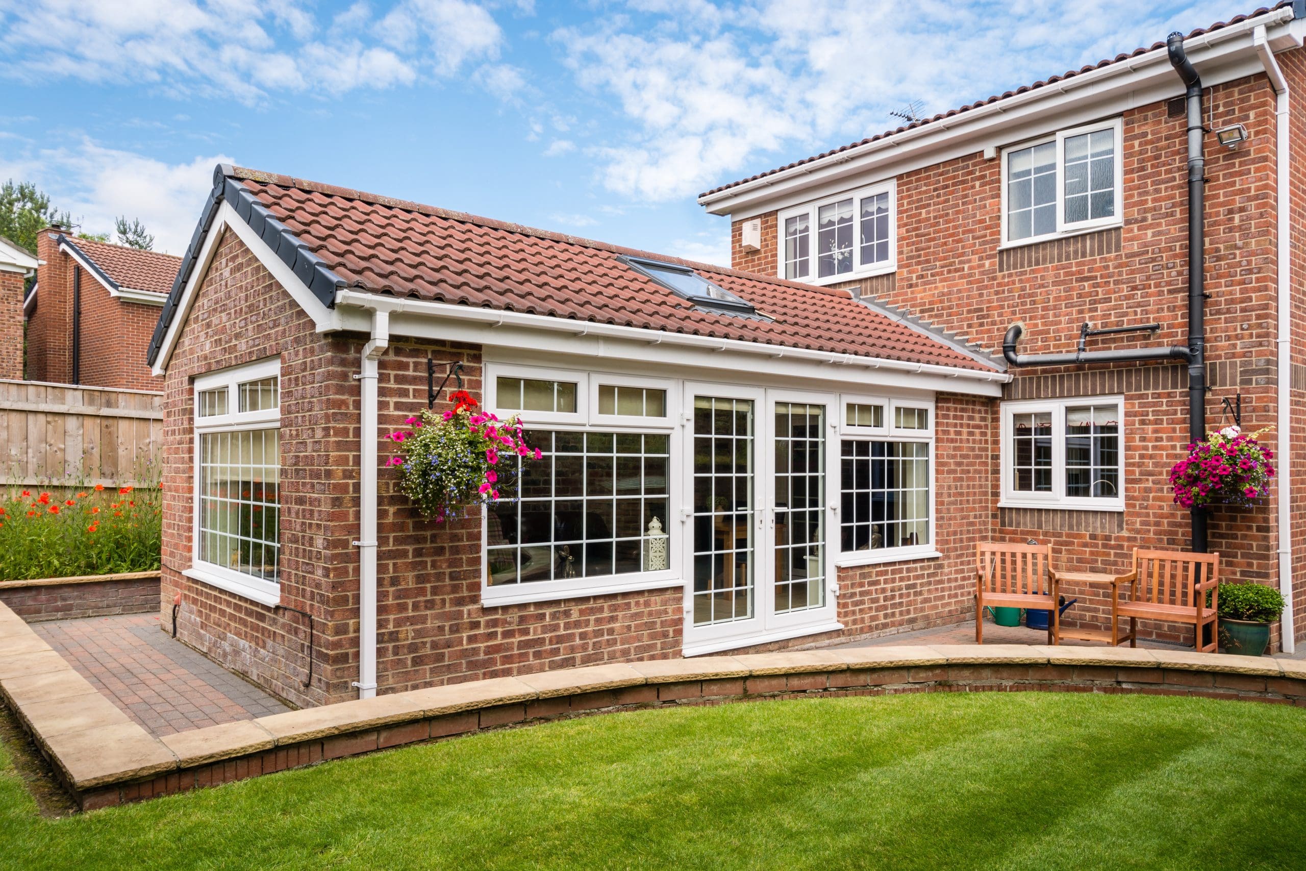 A charming brick house with a red-tiled roof and a sunroom extension is visible. It features large windows and glass doors. Hanging flower baskets and a patio with benches enhance the exterior. A well-manicured lawn completes the scene.