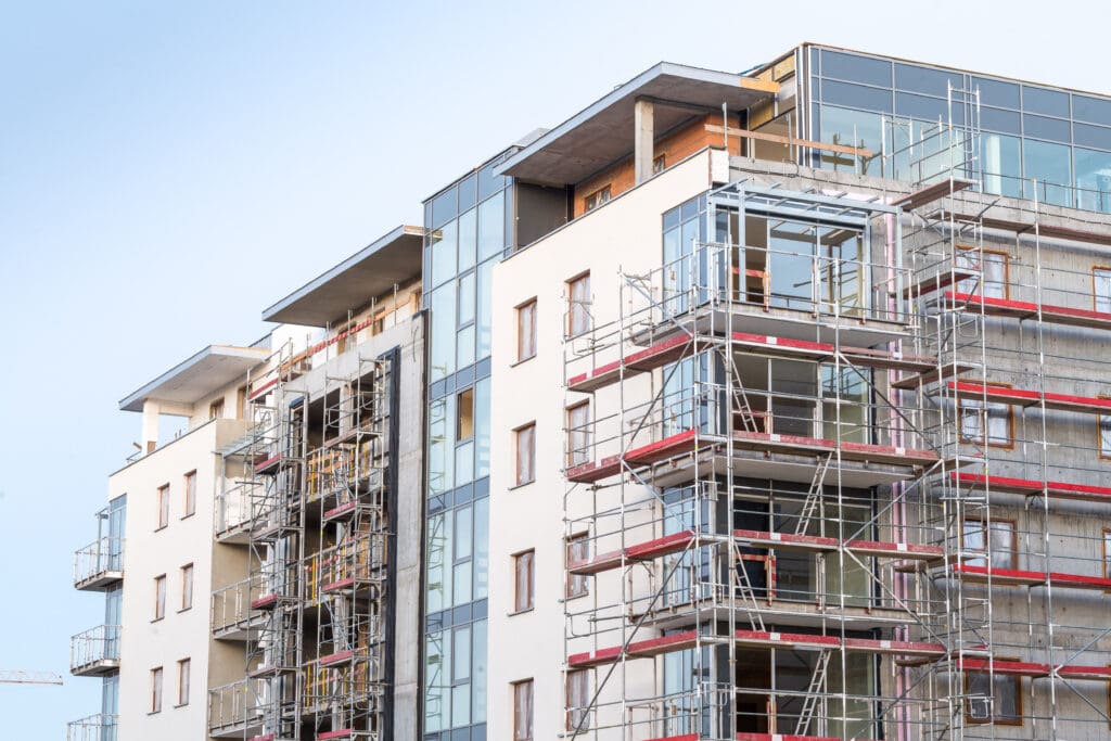 A modern multi-story apartment building under construction, featuring scaffolding on the exterior. The building has large windows and balconies. The sky is clear, suggesting a sunny day.