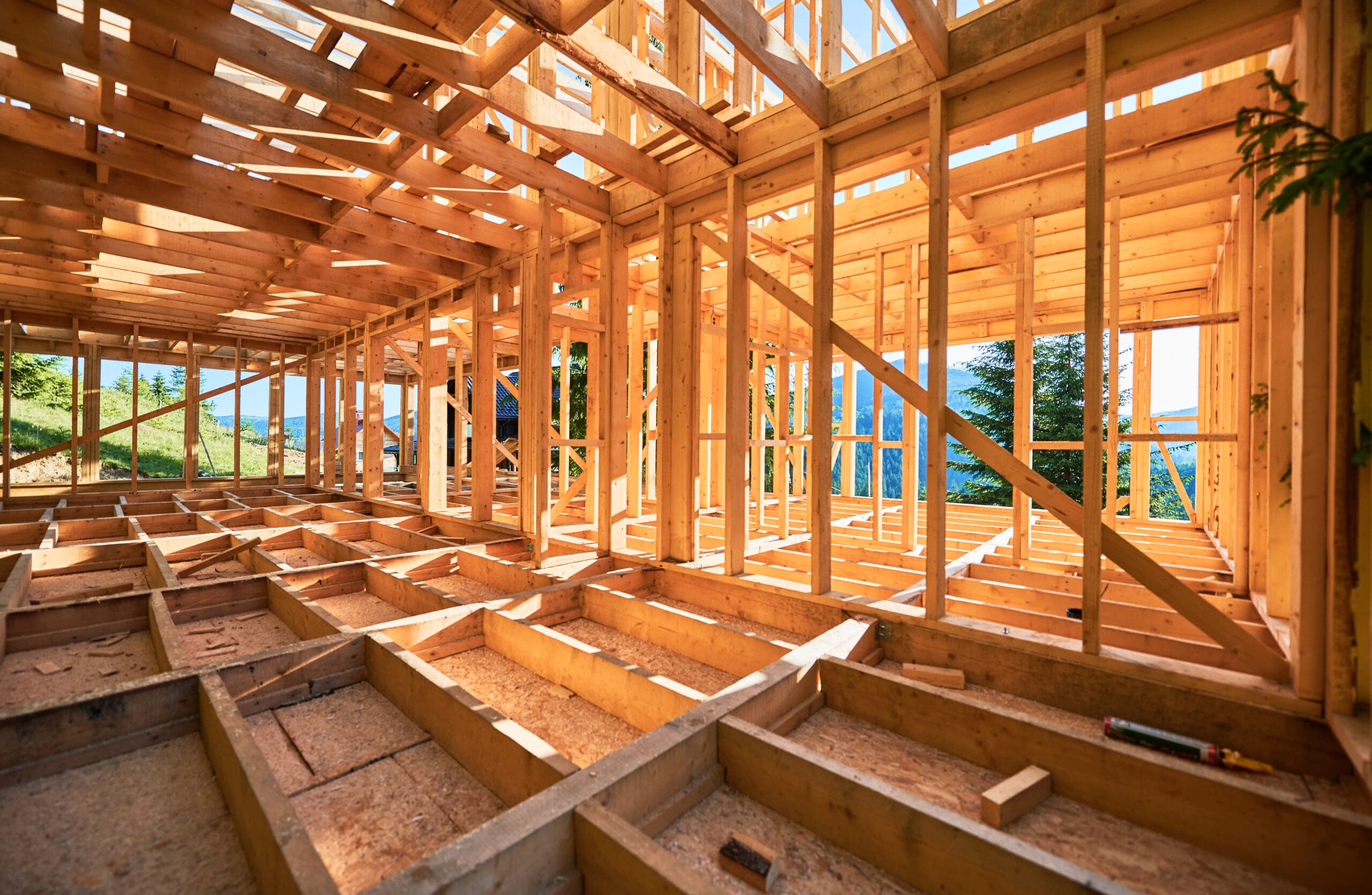 Wooden framing of a house under construction, with visible beams and supports. Sunlight filters through, casting shadows. The background shows a glimpse of greenery and blue sky through the unfinished walls.