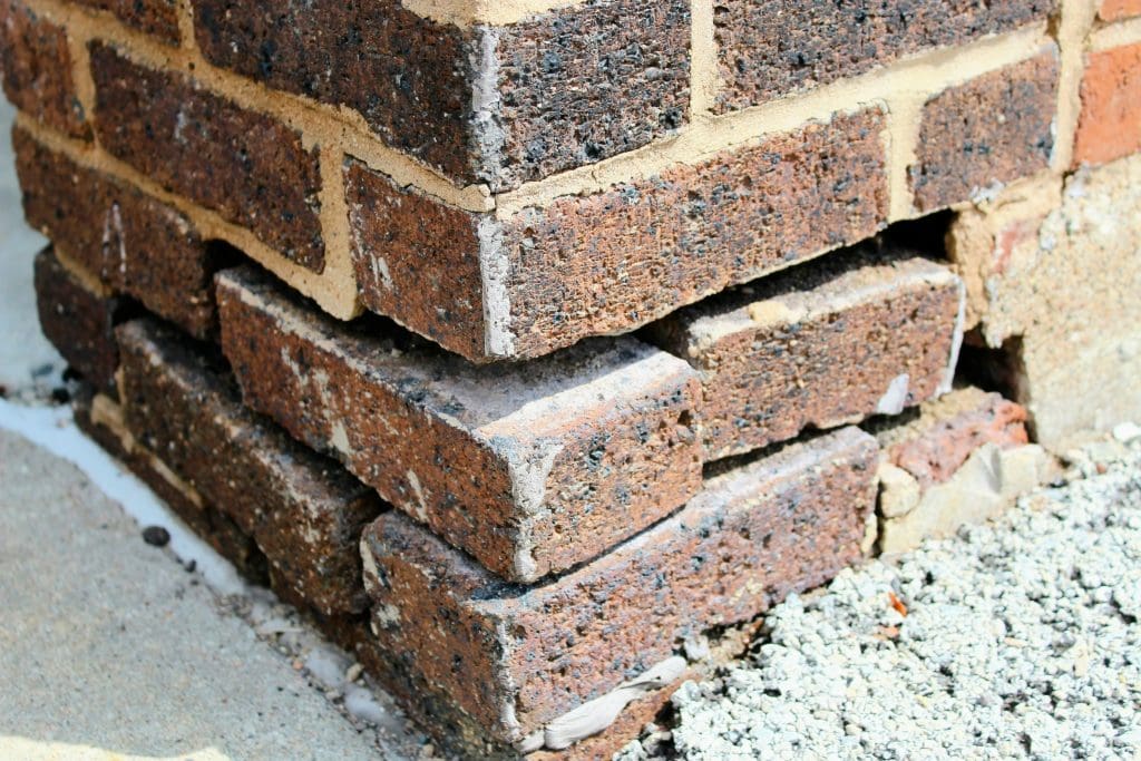 Close-up of a brick wall corner with a section of bricks out of alignment, creating gaps. The bricks are weathered and the mortar is cracked, highlighting damage and structural issues. The ground is made of concrete and gravel.