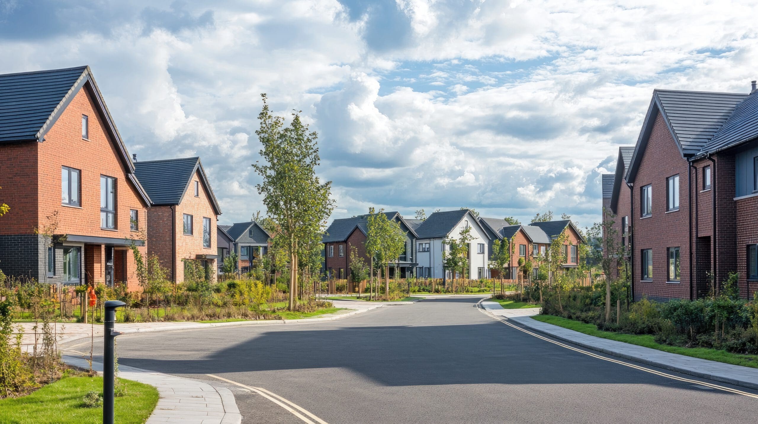 A neighborhood with modern brick houses and manicured lawns under a partly cloudy sky. The curved road is lined with young trees and shrubbery, creating a serene suburban setting.