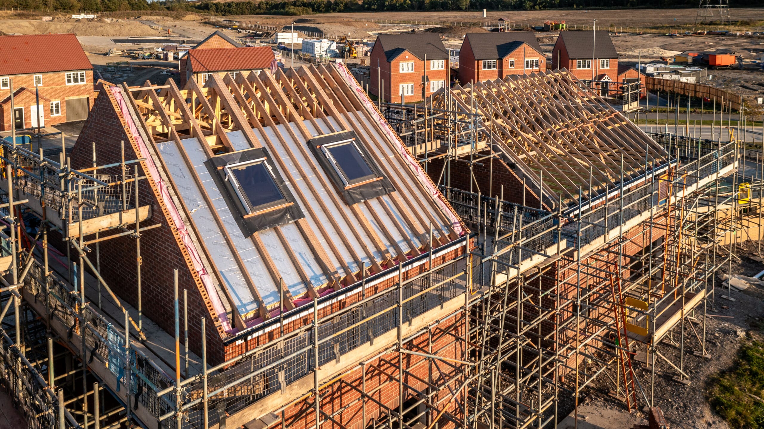 Aerial view of a residential construction site with two houses in the process of having their roofs installed. Scaffoldings surround the buildings, and in the background, finished houses are visible. The area is under development with open grounds.