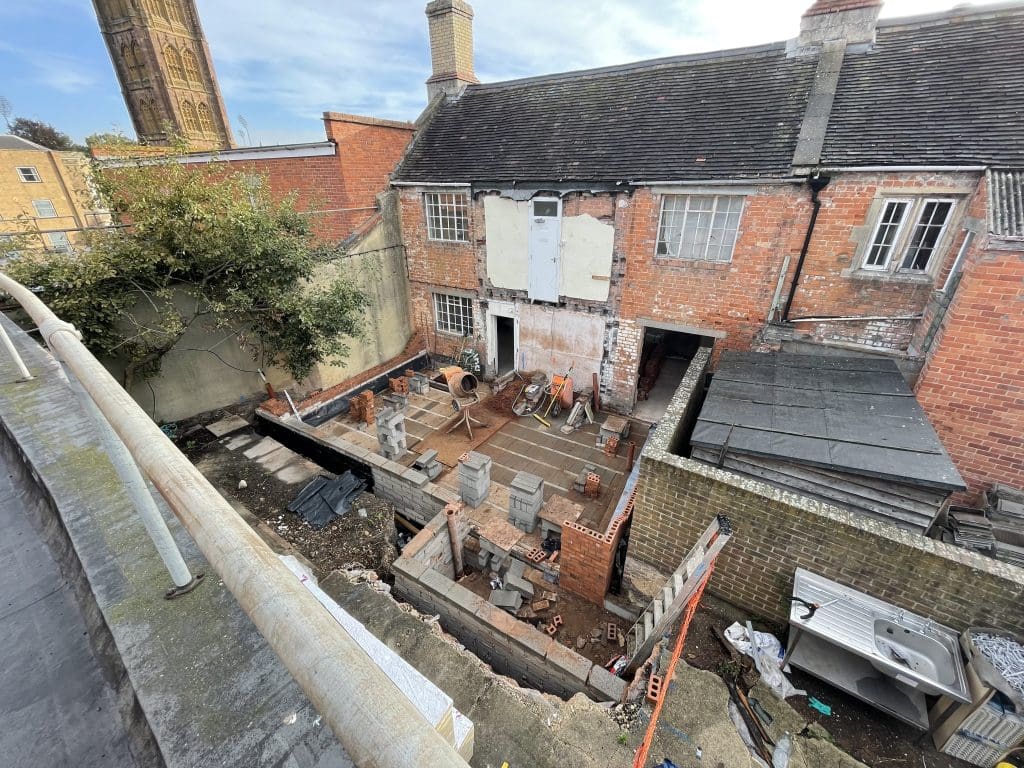 A partially constructed brick building in a small courtyard with construction materials scattered around. The site is surrounded by brick walls and adjacent to older, weathered buildings. A tall tower can be seen in the background.