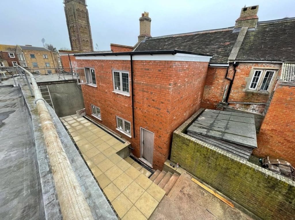 View from a roof overlooking a red brick building with white-framed windows and a sloped roof. A paved area and steps are visible outside. In the background, there are more buildings and a tall tower. The sky is overcast.