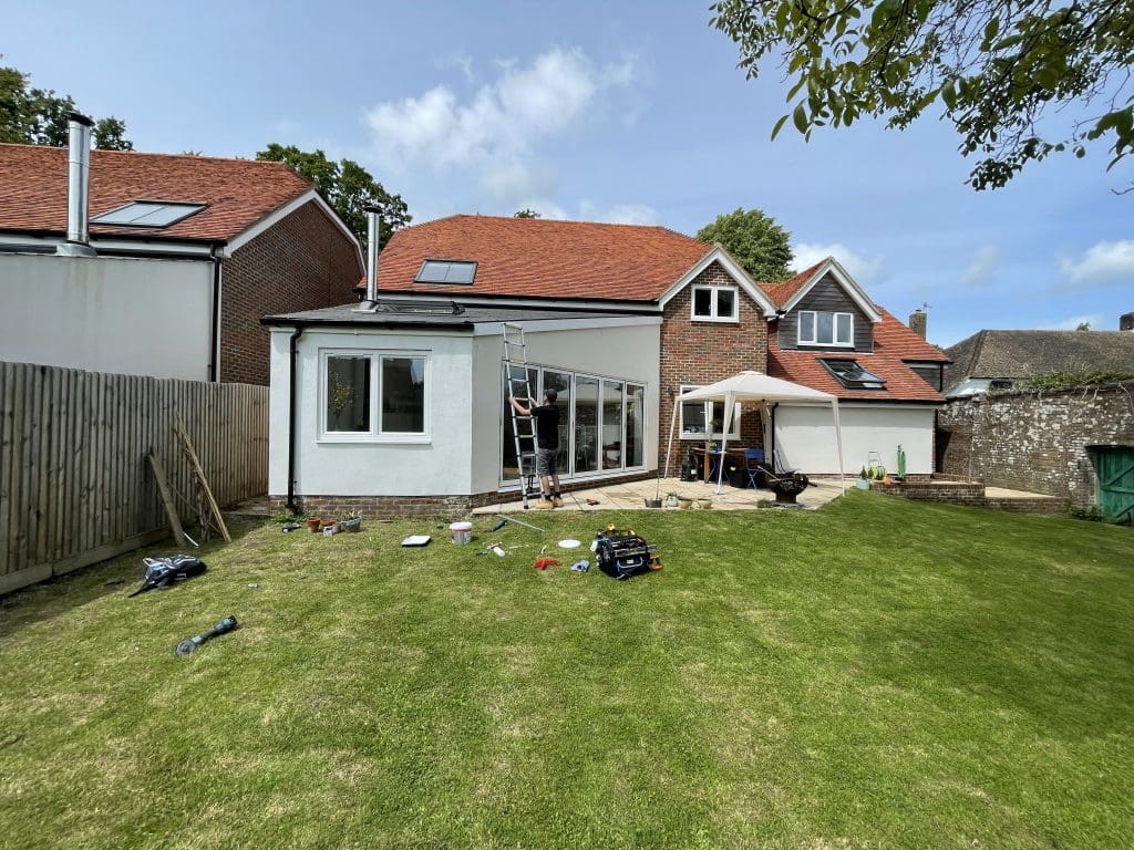 A person stands on a ladder adjusting a satellite dish on a white and brick house with a red roof. The backyard has green grass and a patio area with a canopy and furniture. Various tools and objects are scattered on the lawn.