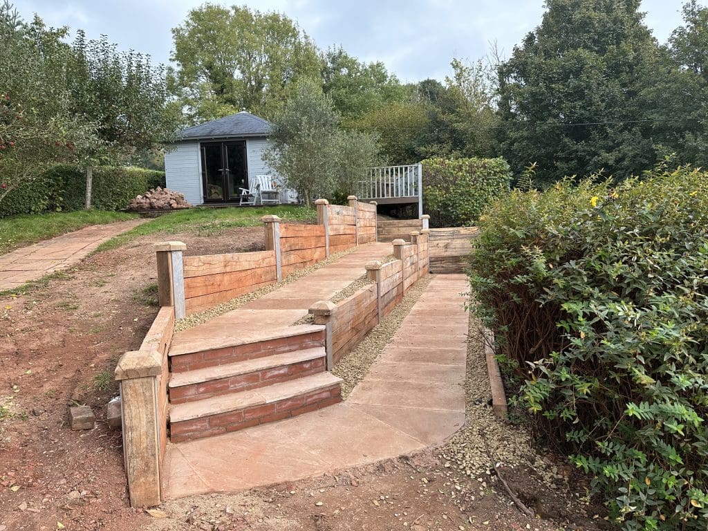 A winding garden path with brown stone steps and railings leads up to a small white cabin surrounded by trees and bushes. The path is bordered by gravel and greenery, with the cabin situated in a tranquil setting.