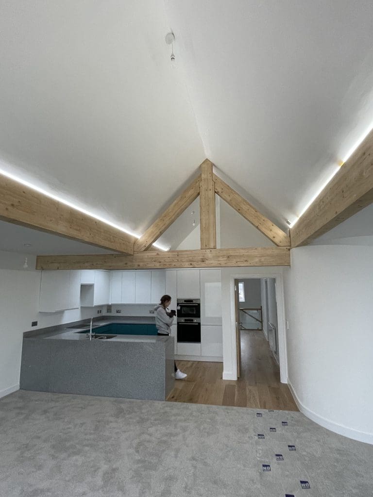 A modern kitchen with exposed wooden beams on the ceiling. A person stands near the center island, which is gray with a built-in sink. The kitchen has white cabinets, and hardwood flooring is adjacent to a carpeted area.