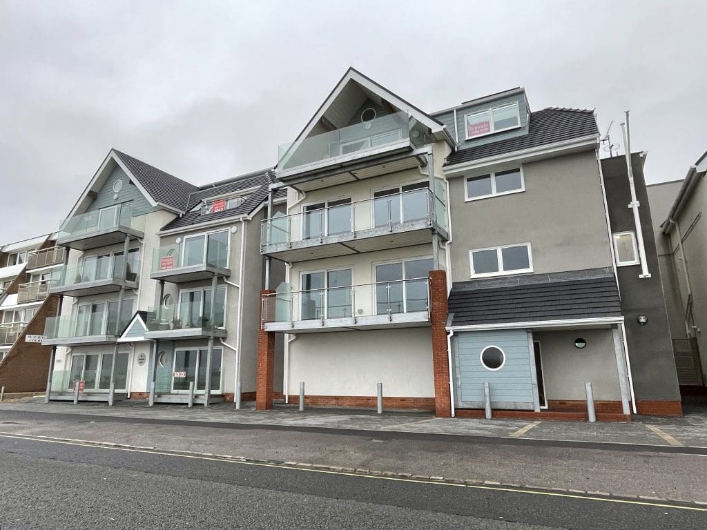 Modern apartment complex with three- and four-story sections. Each unit has glass balconies, and the building features sloped roofs with dormer windows. Signs in some windows indicate availability. The structure sits beside a paved street.