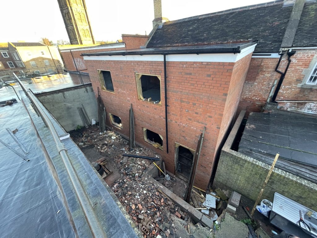 The image shows a partially constructed brick building with window openings and no glass. The area around the building is cluttered with construction debris. There's a visible church tower in the background and adjacent rooftops.