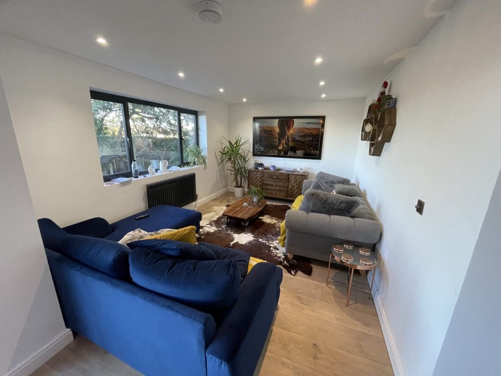 A modern living room with a blue sofa and gray loveseat, a wooden coffee table, and a wall-mounted TV. A large window lets in natural light, and there's a cowhide rug on the floor. Plants and decorative items are placed throughout the room.