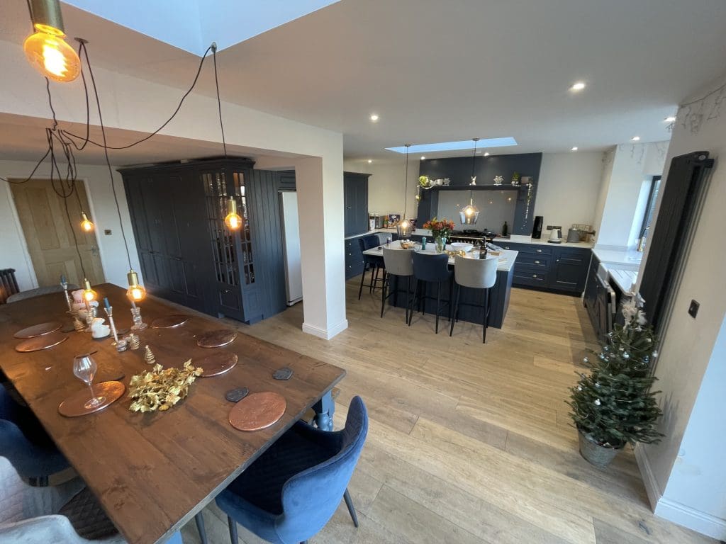 Modern open-plan kitchen and dining area with wooden floors. Features include a wooden dining table with copper place settings, navy chairs, pendant lights, a kitchen island with chairs, and festive decorations, including a small Christmas tree.