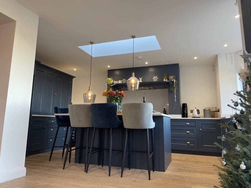 A modern kitchen with dark blue cabinets and wooden flooring. A central island features three upholstered bar stools and two hanging pendant lights. A skylight brightens the space, and a Christmas tree is partially visible in the corner.