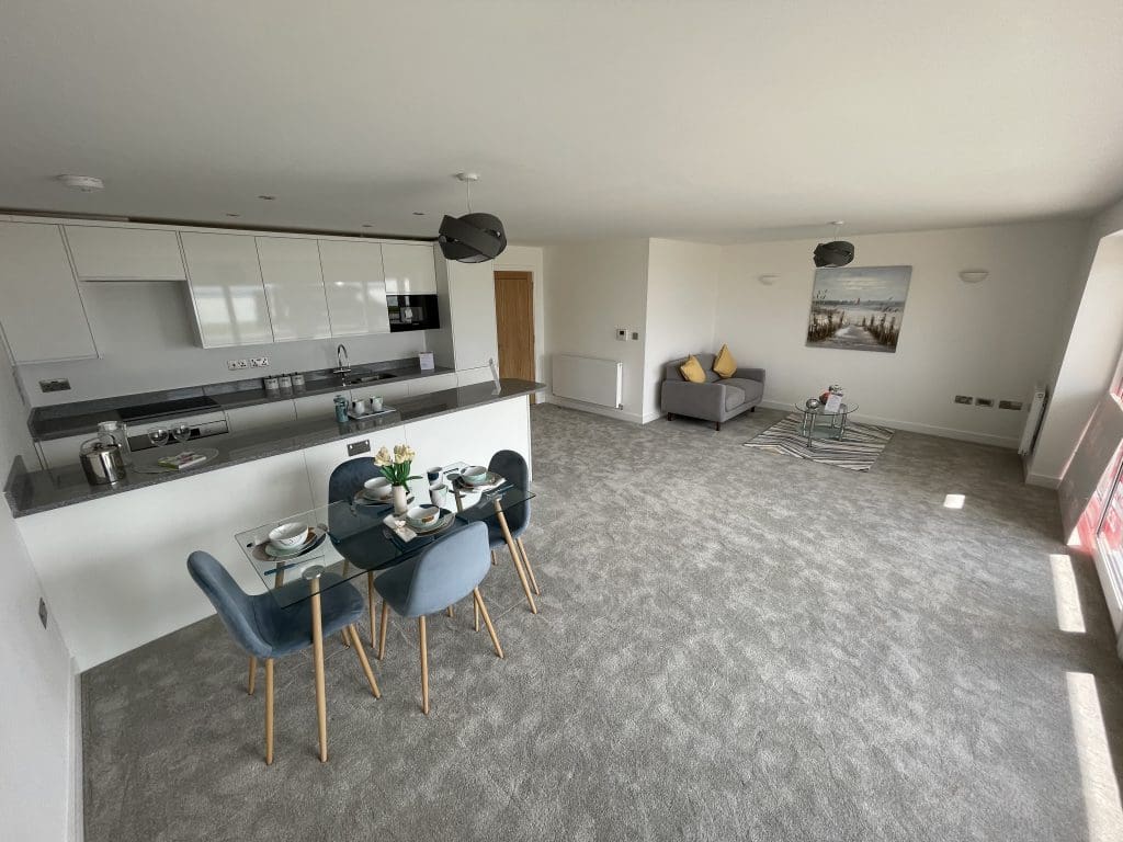 Open-plan living room and kitchen with modern decor. Grey carpet, a dining table with blue chairs, and a sofa with cushions. A painting hangs on the wall, and large windows bring in natural light. White cabinetry and a sleek countertop complete the kitchen.