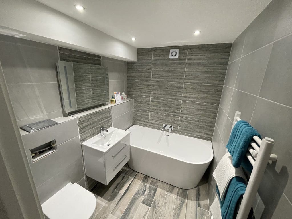 A modern bathroom with grey tiles features a sleek white bathtub, a floating vanity with a sink, a wall-mounted mirror, a toilet, and a towel rack with blue and white towels. The room is well-lit with ceiling spotlights.