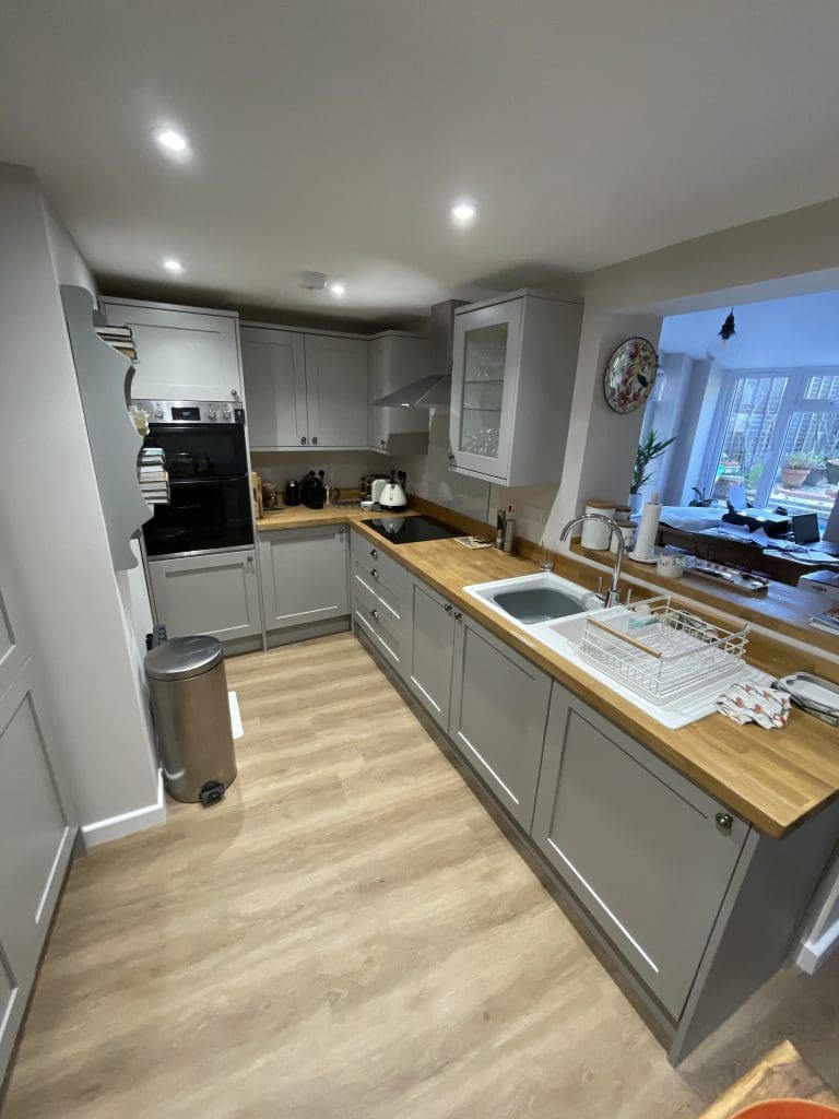 Modern kitchen with gray cabinetry, wooden countertops, and a stainless steel oven and bin. A white sink and dish rack are on the right. The room is well-lit, with a view into a sunlit dining area. Light wood flooring complements the space.