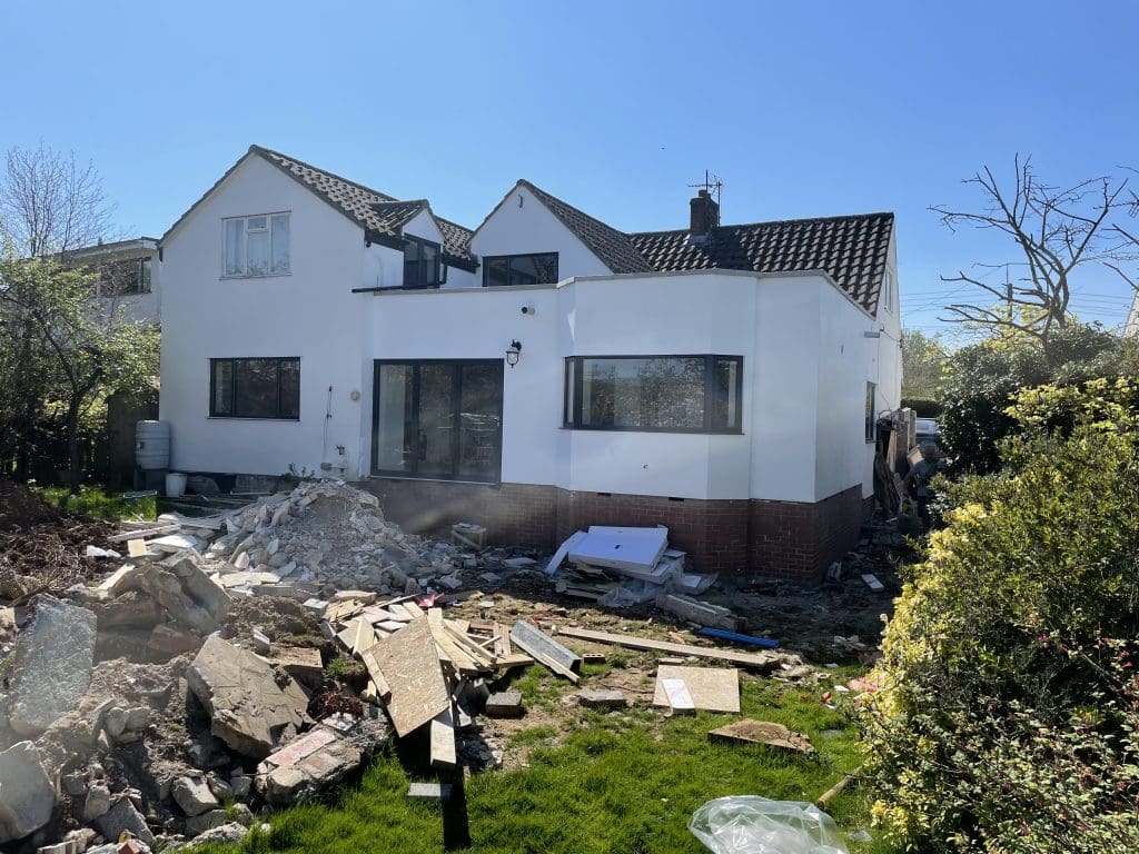 A white two-story house with a black roof is under renovation. Debris and construction materials are scattered on the lawn. Trees and shrubs surround the area under a clear blue sky.