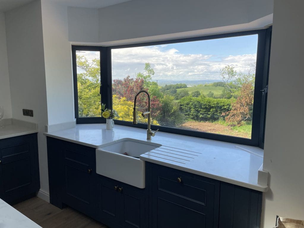 Modern kitchen with dark cabinets and a white countertop featuring a farmhouse sink. A large window offers a scenic view of a green landscape with trees and cloudy sky, creating a bright and airy atmosphere.