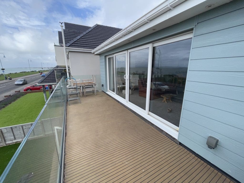 A rooftop terrace with wooden decking and glass railing overlooks a coastal road. The terrace features outdoor furniture, and the building has light blue siding with large sliding glass doors leading inside. The sky is partly cloudy.