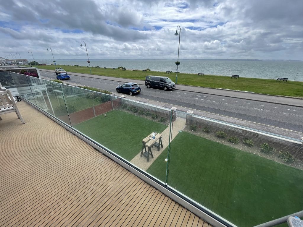 View from a balcony with glass railing overlooking a road, grassy area, and the sea. Three vehicles are on the road, with a cloudy sky above. Small table and chairs on the grass below.