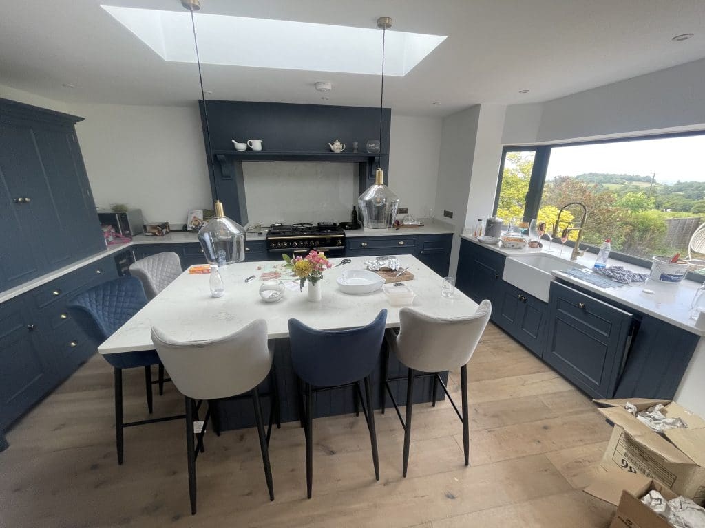 A modern kitchen with blue cabinets and a large central island. There are five chairs around the island, a range oven, and two pendant lights above. A skylight and large window offer natural light. A vase with flowers and various kitchen items are on the countertop.