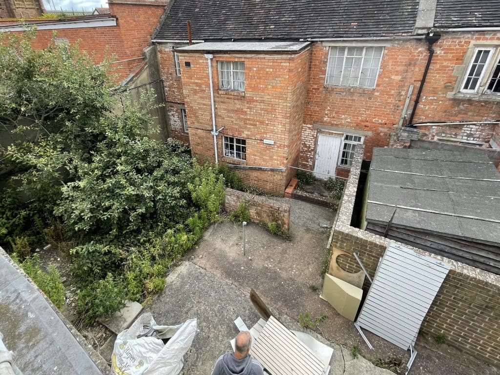 A person stands in a small, overgrown courtyard surrounded by brick buildings and wooden fences. Various building materials and debris are scattered around the courtyard. The scene is viewed from an elevated angle.