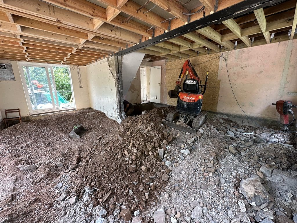 A construction site inside a building with exposed wooden beams. A small excavator is digging into a pile of dirt and rubble. Partially demolished walls and a large glass door leading outside are visible.