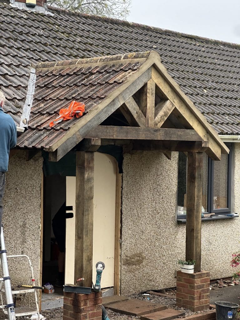 A construction worker on a ladder installs a wooden porch frame with a tiled roof over a doorway of a pebble-dashed house. A small potted plant is on a brick pier supporting the porch. A few tools and construction materials are scattered around.