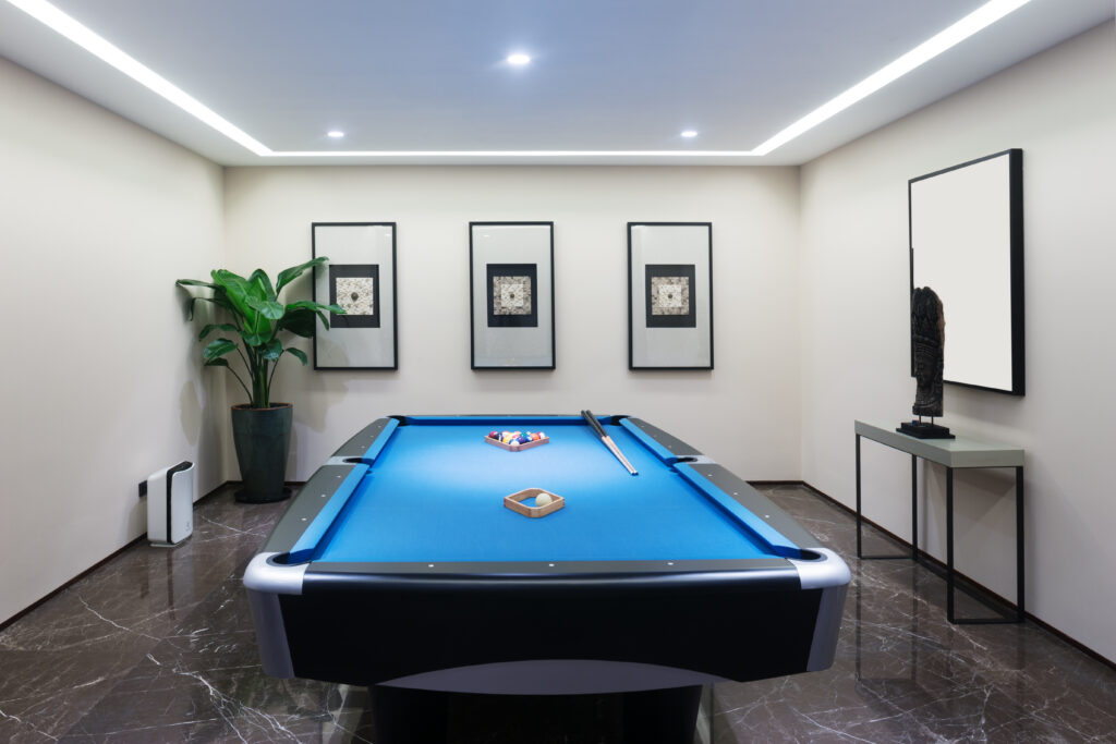 A modern game room with a blue pool table, cue sticks, and balls set up. The space has marble flooring, a large potted plant, framed wall art, and a sculptural figure on a side table.