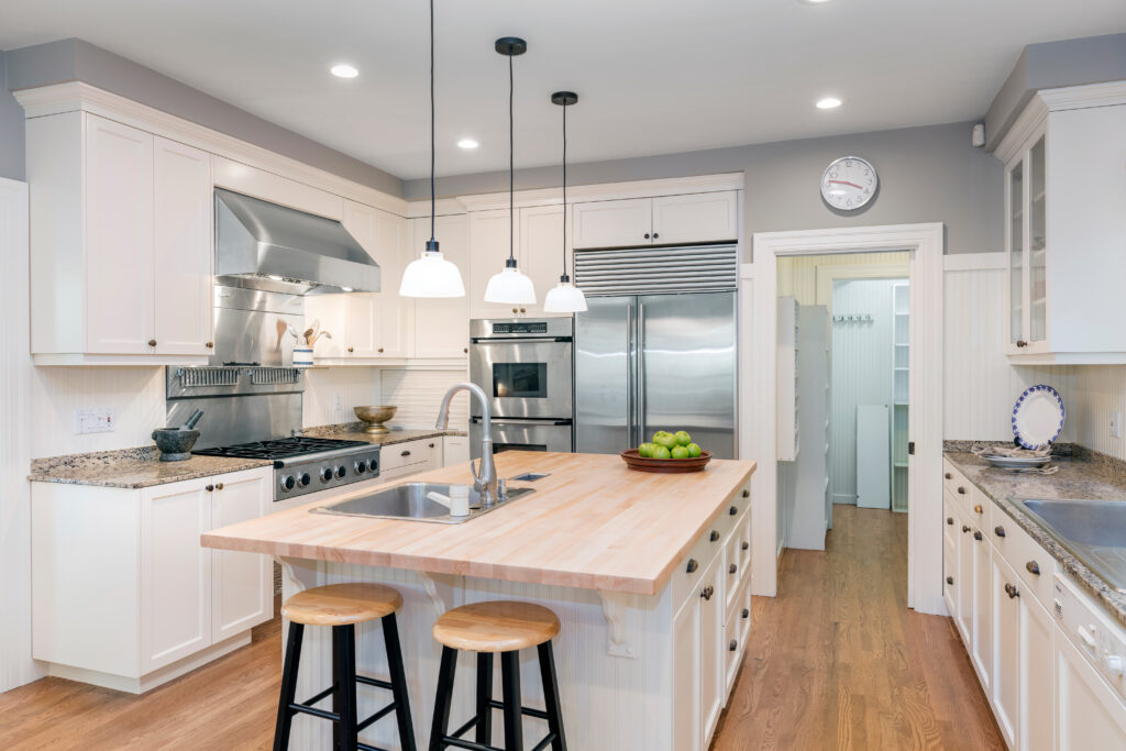 Bright, modern kitchen with white cabinets, stainless steel appliances, a wooden island with sink, two wooden stools, hanging lights, and a bowl of green apples. Doorway leads to a pantry with shelves.
