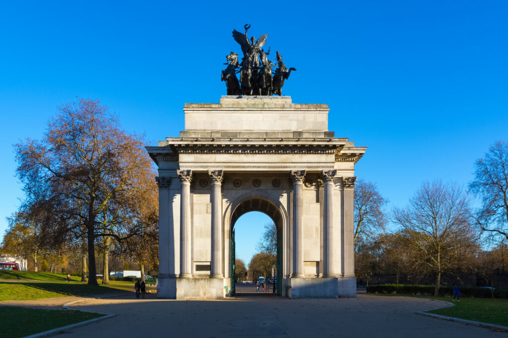 The photo shows the Wellington Arch in London, a large stone arch with a bronze quadriga statue on top, set against a clear blue sky and surrounded by trees.