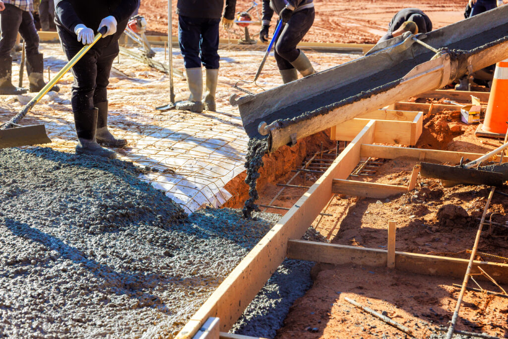 Workers in boots use tools to spread and smooth freshly poured concrete from a chute onto a rebar-framed foundation at a construction site.