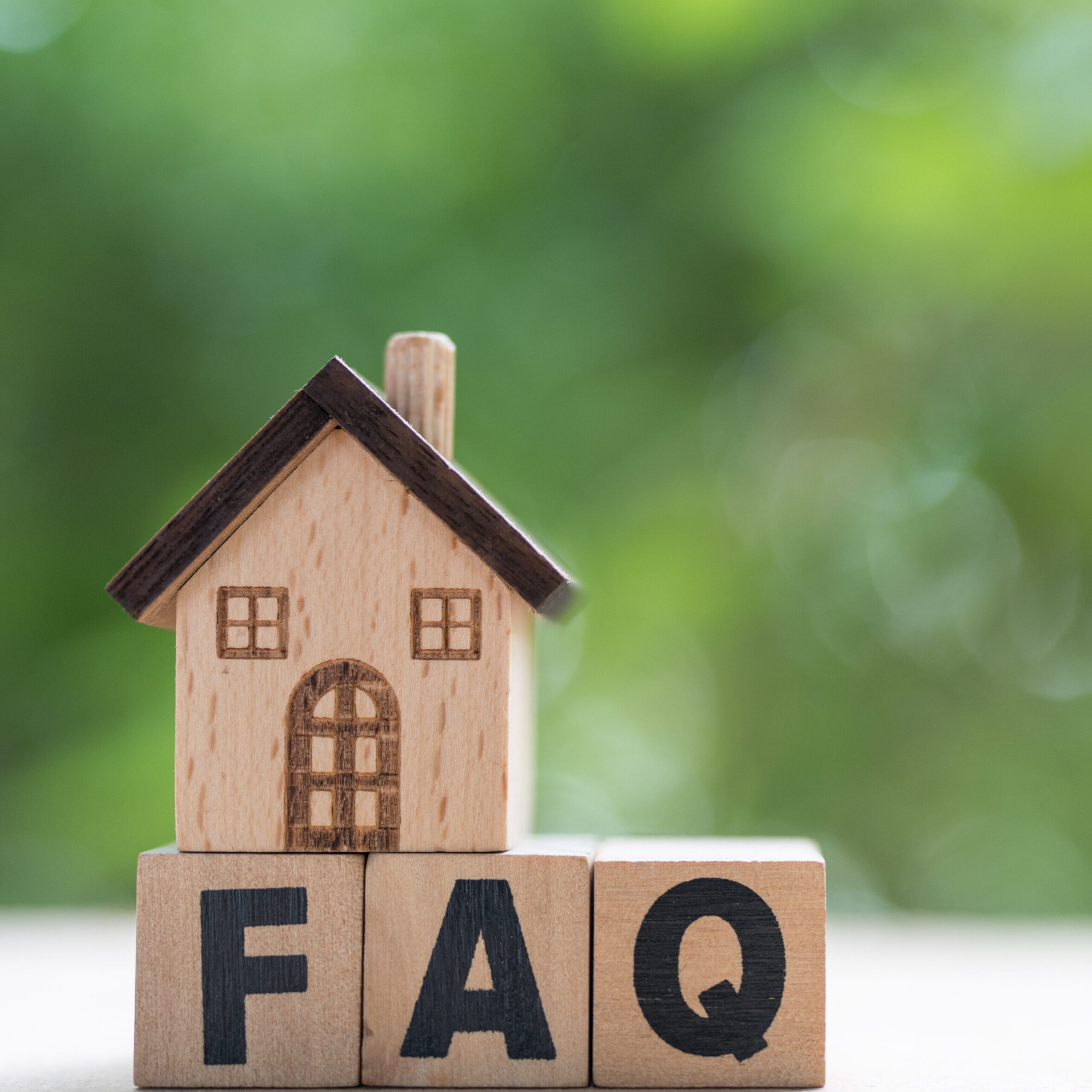 A small wooden house model sits on wooden blocks that spell out "FAQ" against a blurred green background.