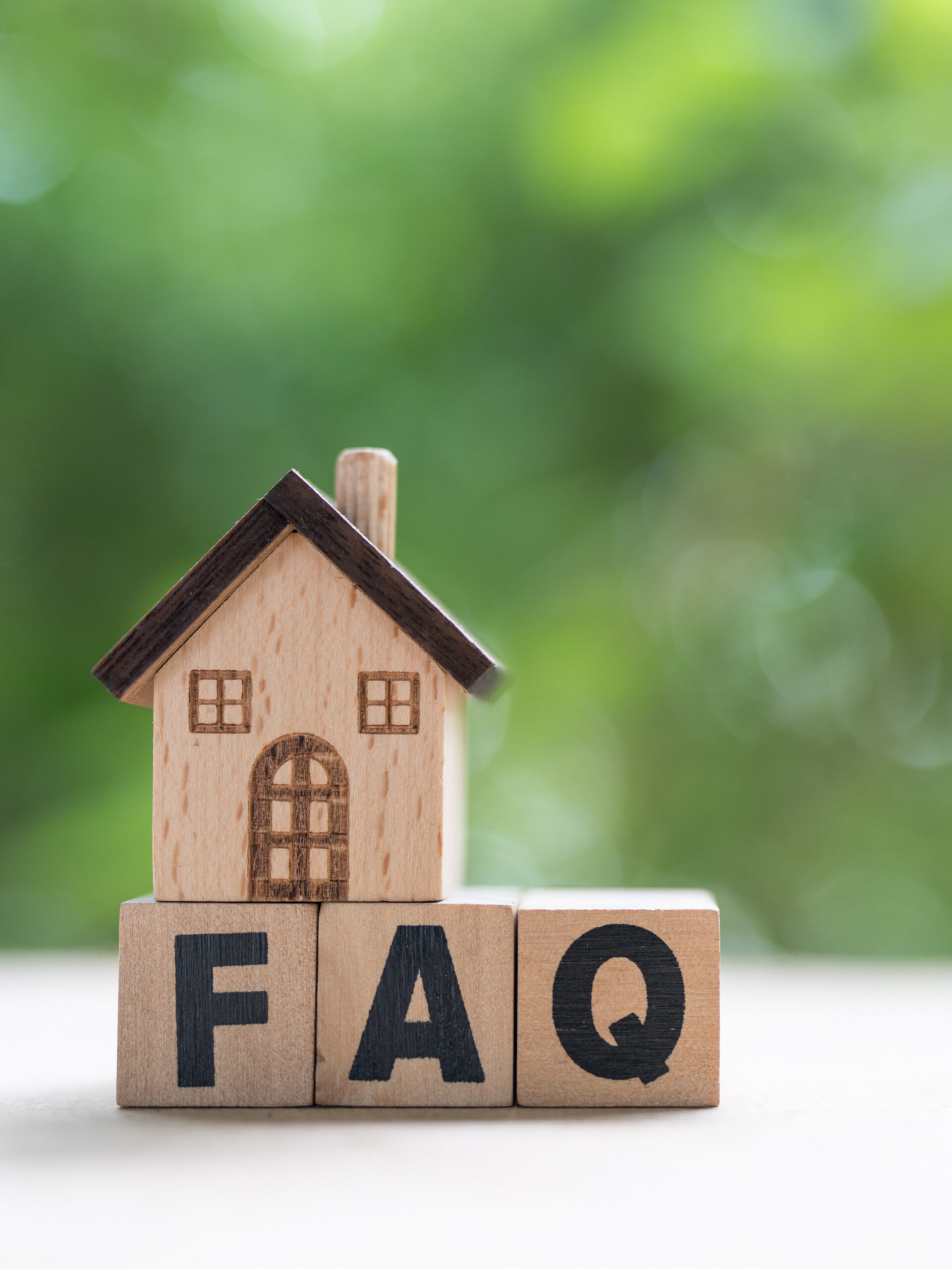A small wooden house model sits on top of three wooden blocks that spell "FAQ," with a blurred green background.