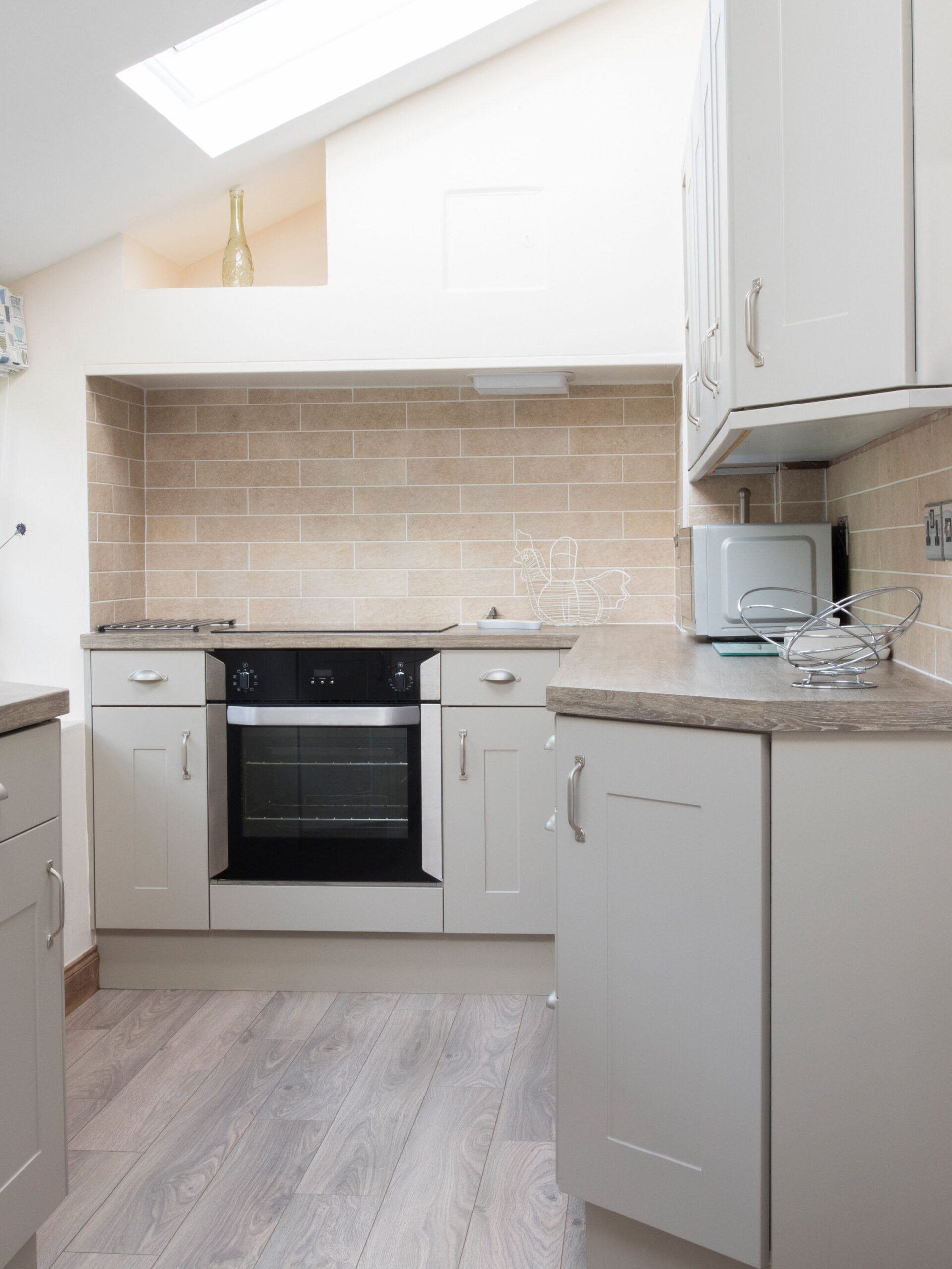 A small, modern kitchen with light gray cabinets, a black oven, and a beige tile backsplash. Natural light enters through a skylight. There is a microwave on the counter and a stainless steel sink with a drying rack, near a window.