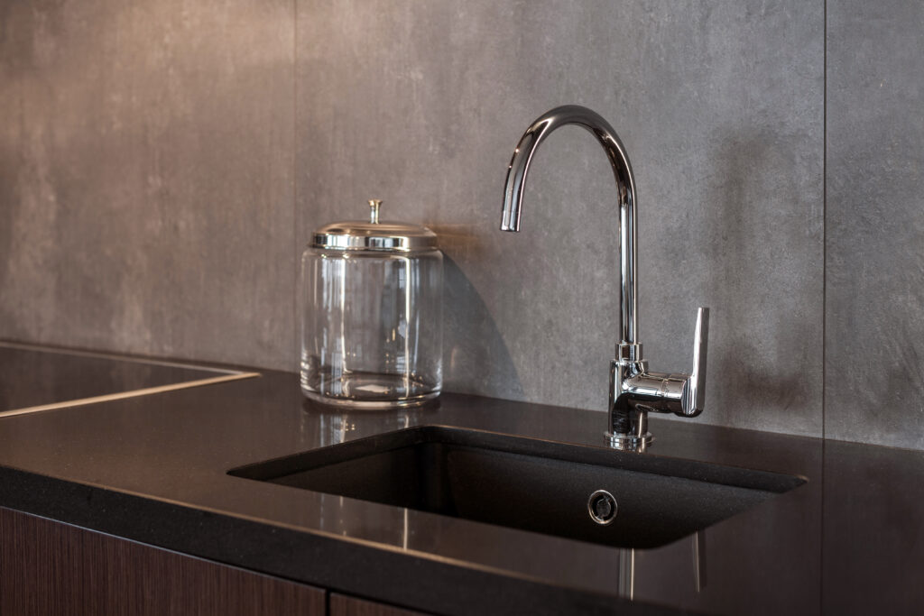 A modern kitchen sink with a sleek chrome faucet, set in a dark countertop. A clear glass jar with a metallic lid sits next to the sink. The background features a gray textured wall.