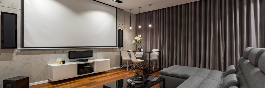 Modern home theater room with a large projector screen, wall-mounted speakers, a white media console, a gray sectional sofa, a small dining table with white chairs, and floor-to-ceiling gray curtains.